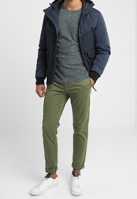Giacca a felpa navy con una camicia grigia testurizzata, pantaloni verde oliva e sneakers bianche; presenta tasche con zip e polsini elasticizzati.