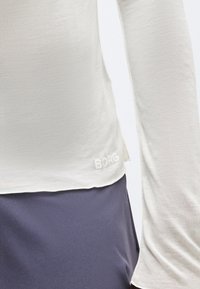 Κοντινή λήψη ενός ατόμου που φοράει κρεμ πουκάμισο με μακριά μανίκια, με την επιγραφή "BORG" κοντά στο κάτω μέρος και σκούρα φούστα ή παντελόνι.