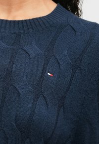 Granatowy sweter w splot warkoczowy z żebrowanym okrągłym dekoltem oraz małym haftowanym logo w kolorach czerwonym, białym i niebieskim z przodu.