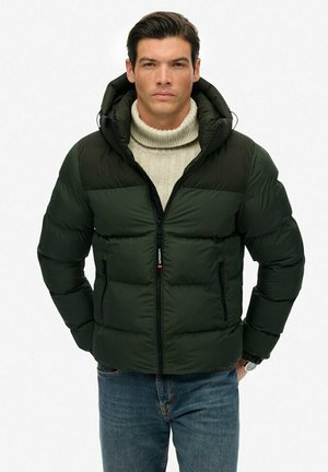 Hombre vistiendo una chaqueta acolchada verde oscuro sobre un suéter de cuello alto color crema y jeans azules, de pie con las manos en los bolsillos de la chaqueta.