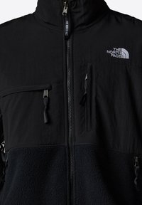 Black jacket featuring a mix of smooth fabric and fleece texture. Zippered pockets on the chest and left side. Brand logo stitched in white.