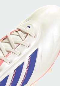 Botas de fútbol sintéticas blancas con acentos rosas, que presentan rayas azules y una superficie texturizada; diseño con cordones; tacos moldeados para tracción.