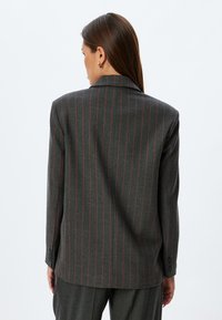 Blazer gris rayé avec des lignes rouges, design structuré, devant simple boutonnage et poignets à deux boutons. Tissu lisse avec une coupe ajustée.