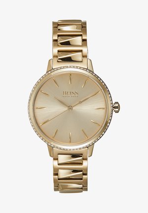 Reloj de pulsera Hugo Boss dorado con esfera redonda, marcadores de hora dorados minimalistas, manecillas delgadas y una correa metálica enlazada con detalles de cristales.