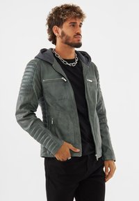 Giacca in pelle da uomo di colore verde tenue con dettagli trapuntati sulle spalle, cerniere argentate e cappuccio grigio. Una camicia e pantaloni neri completano l'outfit.