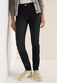 Jeans skinny negros de denim, con un diseño clásico de cinco bolsillos y cierre de botón frontal, combinados con zapatillas de colores claros.