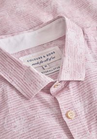 Camicetta a maniche corte di colore rosa chiaro con righe bianche e rosse, dotata di colletto button-down e una patta con un bottone. Etichetta visibile.