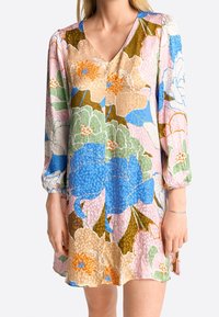 Nainen pukeutuneena pitkähihaiseen, V-päänteellä varustettuun mekkoon, jossa on suuri monivärinen kukkapainatus sinisen, vaaleanpunaisen, vihreän ja beigensävyjen yhdistelmässä.