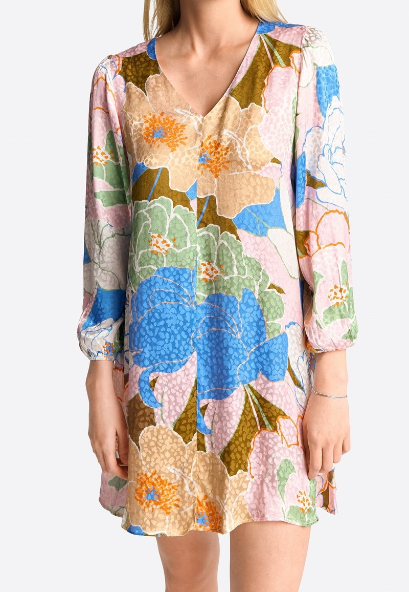 Nainen pukeutuneena pitkähihaiseen, V-päänteellä varustettuun mekkoon, jossa on suuri monivärinen kukkapainatus sinisen, vaaleanpunaisen, vihreän ja beigensävyjen yhdistelmässä.