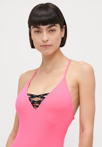 Un maillot de bain rose avec de fines bretelles, présentant un décolleté en V profond et des détails en croix noirs avec des logos. Le matériau semble lisse et ajusté.