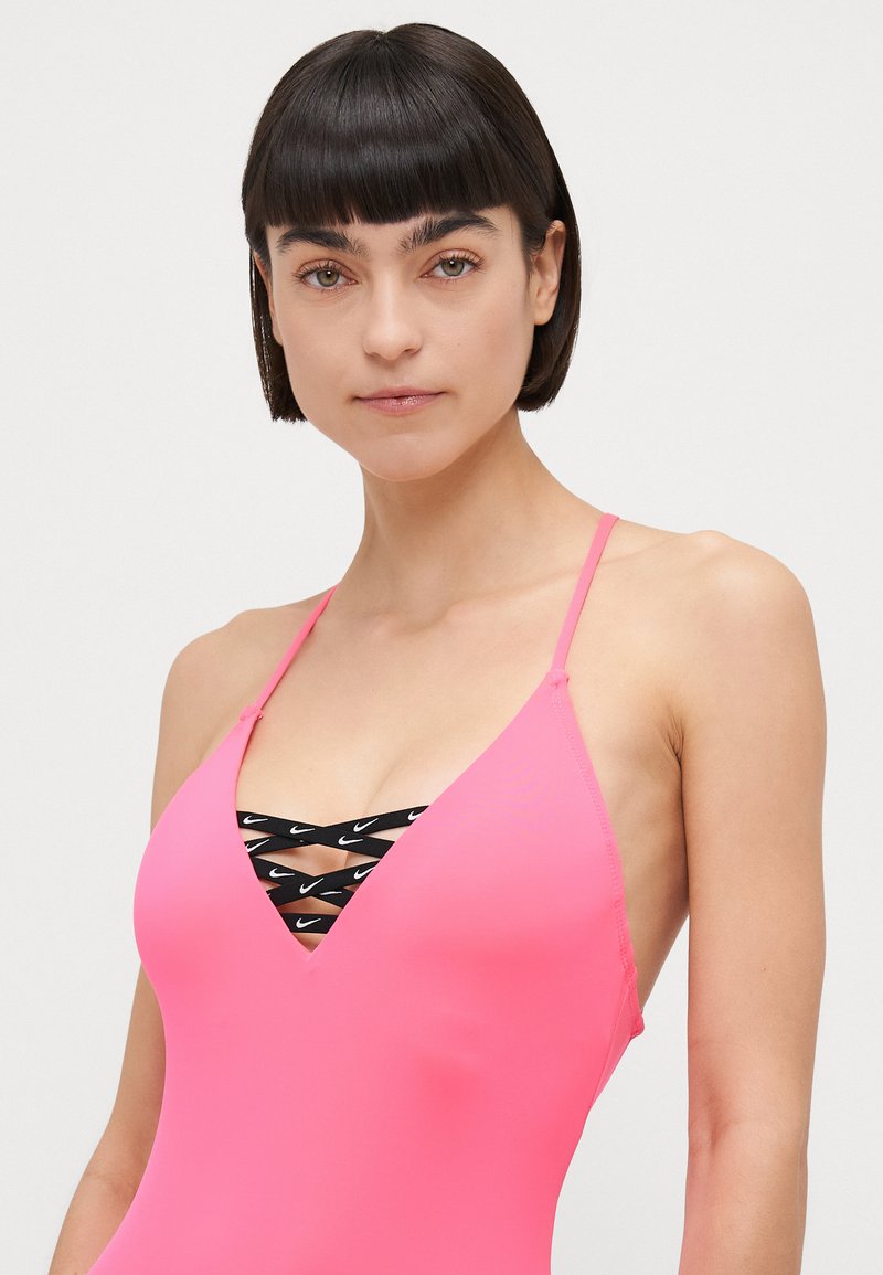 Costume da bagno rosa con sottili spalline, presenta una profonda scollatura a V e dettagli incrociati neri con loghi. Il materiale appare liscio e aderente.