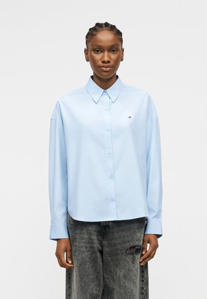 Kobieta z zaplecionymi włosami nosi jasnoniebieską koszulę na guziki i wyblakłe czarne dżinsy z rozdarciem na prawej udzie, stoi na białym tle.