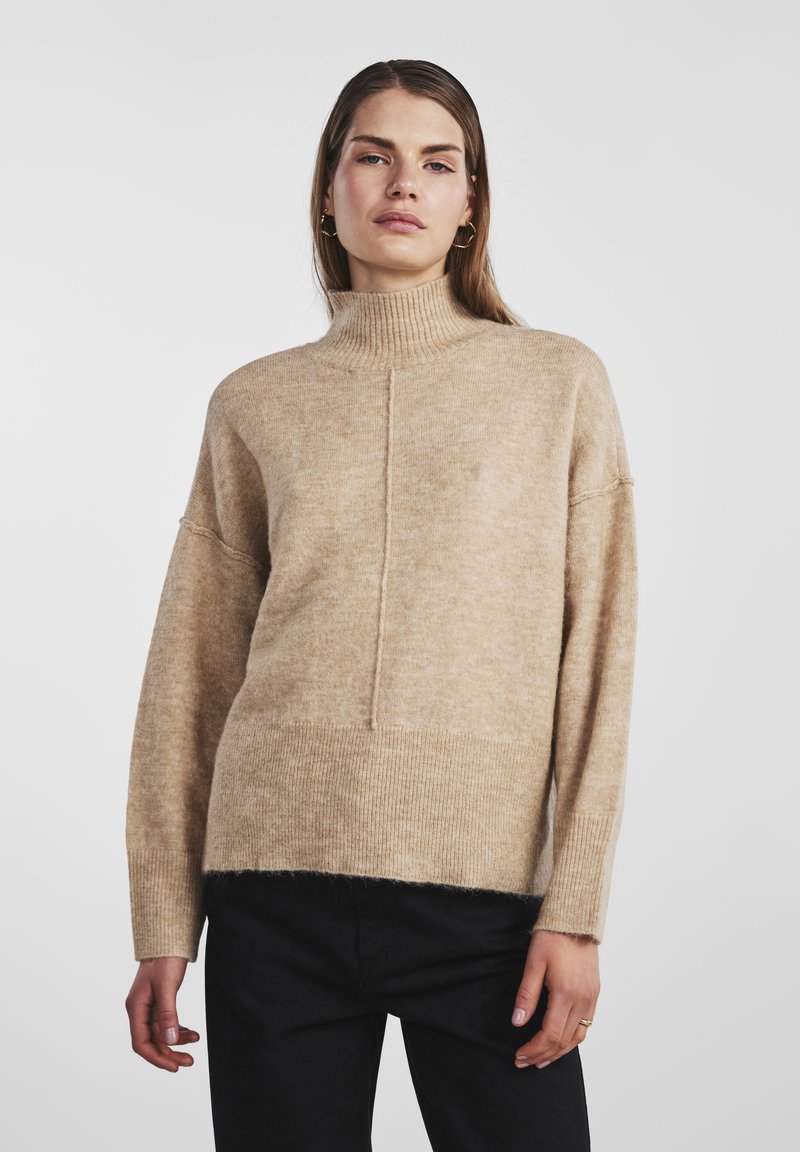 Bež jopica s turtleneck izrezom z mehak, teksturiran zaključek, ki ima ravno silhueto, spuščena ramena ter rebraste robove in manšete.