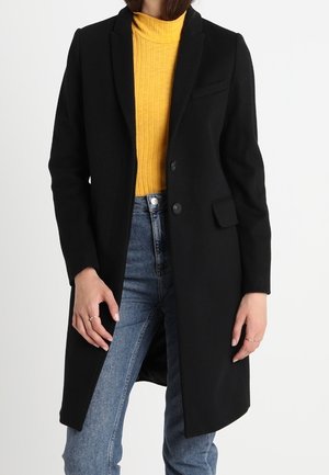 Kvinde iført en sort lang frakke over en gul ribstrikket rullekrave og blå jeans, stående mod en ensartet hvid baggrund.