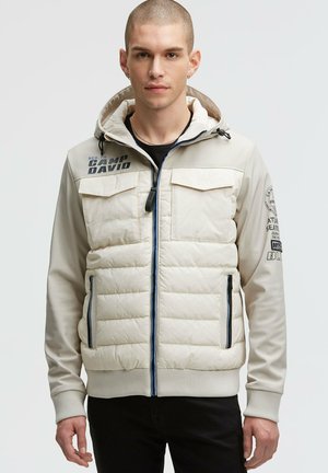 Beige puffer jacket with a hood, featuring blue zipper, side pockets, and ribbed cuffs. Includes printed text and logo details on the front.