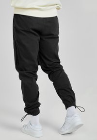 Person i svarta cargo-byxor med justerbara ankelband, vita strumpor och vita Nike-sneakers mot en enkel ljus bakgrund.