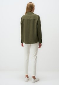 Olijfgroene shirt met een kraag, knoopmanchetten en een ronde zoom; gecombineerd met witte broek en lichte sneakers; gladde textuur.