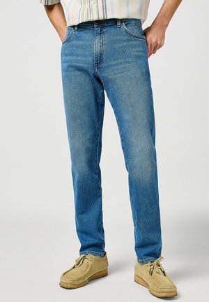 Light blue denim jeans with a straight cut, featuring five pockets and a faded finish, worn with beige suede shoes.