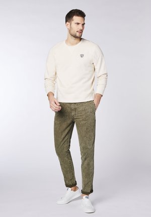Beige Sweatshirt mit runder Ausschnitt und kleinem Bärenlogo auf der Brust, kombiniert mit olivgrünen, strukturierten Hosen und weißen Turnschuhen.