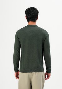 Vyras su tamsiais plaukais, apsirengęs tamsiai žalia ilgos rankovės megztiniu ir smėlio spalvos kelnėmis, atsukęs nugarą prieš baltą vientisą fono.