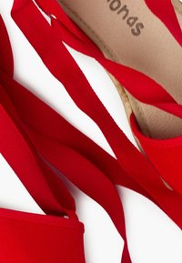 Zapato de tela roja con una amplia correa de cinta, diseño plano y una parte superior texturizada. La plantilla es beige y hay un detalle de costura visible.