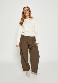 Biele dlhé rukávy s hnedými plisovanými širokými nohavicami. Modelka má na sebe béžové topánky s ostro špičkou. Hladké textúry a jemný farebný kontrast.