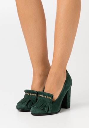 Des chaussures à talons hauts en daim vert avec un devant à franges et des détails en métal. Les talons sont de forme bloc et robustes.