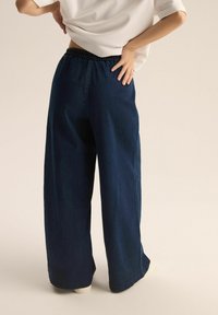 Dżinsowe spodnie z szerokimi nogawkami w ciemnoniebieskim kolorze z elastycznym pasem. Gładka tekstura i czyste szwy. Prosty krój bez widocznych wzorów czy ozdób.