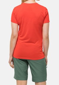 Osoba zobrazená zezadu, oblečená v jednoduchém červeném tričku s krátkým rukávem a zelených kratkých šortkách do kolen na jednotném pozadí.