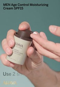 Crema hidratante de control de edad para hombres en un bote beige con una tapa de bomba marrón oscuro, sostenida entre los dedos, que dispensa una pequeña cantidad de crema.
