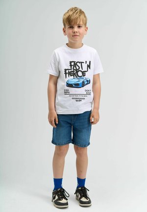 Tricou alb cu imprimeu grafic negru și albastru care prezintă o mașină; asociat cu pantaloni scurți din denim albastri și adidași negri cu accente albe.