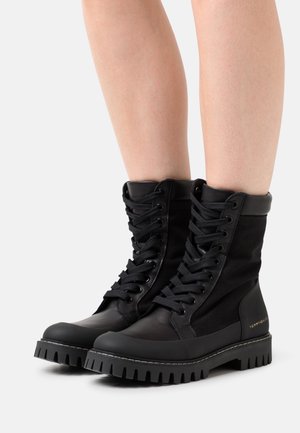Bottes de combat noires en cuir et tissu avec lacets, semelles crantées épaisses, portées sur des jambes nues sur fond blanc.