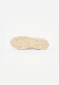 Hvid sneaker sål med en struktureret beige gummibund, der har et bølget mønster og et cirkulært logo ved hælområdet.