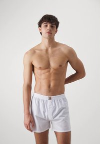 Fehér pamut boxernadrág elasztikus derékrésszel és kis logófolttal. A modell semleges testtartásban áll sima háttér előtt.