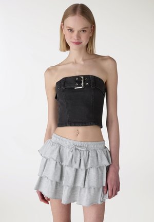 Giovane donna che indossa un top nero senza spalline con fibbia e una minigonna grigia chiara arricciata, in piedi contro uno sfondo semplice.