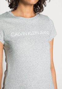 Světle šedé bavlněné tričko s kulatým výstřihem a krátkými rukávy. Na hrudi je bílý nápis "CALVIN KLEIN JEANS". Hladká textura.
