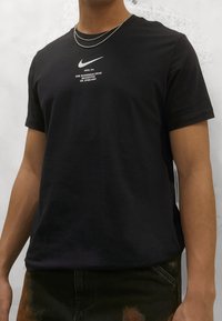 Musta puuvillainen T-paita, jossa on pyöreä kaula-aukko ja lyhyet hihat. Edessä valkoinen Nike-logo ja teksti. Yksinkertainen rakenne ja normaali istuvuus.