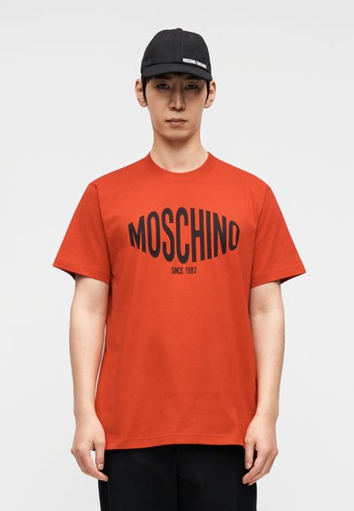Κόκκινο βαμβακερό t-shirt με έντονο μαύρο λογότυπο "MOSCHINO" και κείμενο "SINCE 1983", κοντά μανίκια και στρογγυλή λαιμόκοψη. Φορεμένο με μαύρο καπέλο.