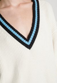 Pull à col V en crème avec une texture côtelée. Présente un col rayé en noir et bleu. Accent sur le matériau tricoté et les couleurs contrastantes.