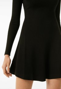 Robe noire côtelée à manches longues, corsage ajusté et jupe évasée. Tissu texturé, finition douce et ourlet lisse au niveau du genou.