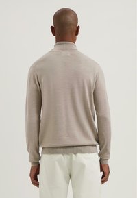 Beige pletený svetr s vysokým l�ímcem, dlouhými rukávy a žebrovanými manžetami, nosený přes bílé kalhoty, z pohledu ze zad.