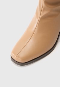Gros plan sur l'orteil d'une botte en cuir beige avec une couture centrale et une semelle marron foncé, présentée sur fond blanc.