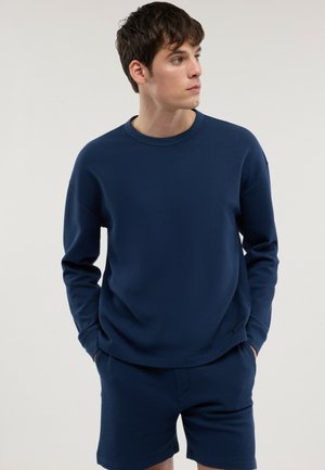 Joven con camisa azul marino texturizada de manga larga y pantalones cortos a juego, mirando hacia su izquierda con las manos en los bolsillos.