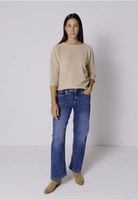 Beige rakennepaita, jossa on kolmen neljänneksen hihat, yhdistettynä sinisiin suoriin farkkuihin. Mallilla on päällään vaaleanruskeat nilkkurit.