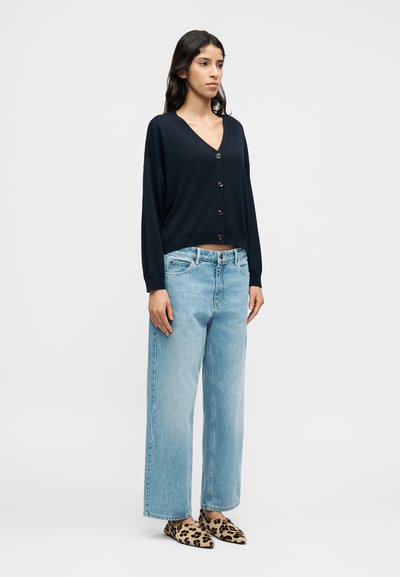 Αμάνικη πλεκτή ζακέτα σε ναυτικό μπλε με V-λαιμόκοψη και τέσσερα κουμπιά, σε συνδυασμό με ανοιχτό μπλε τζιν παντελόνι με φαρδιά μπατζάκια και παπούτσια τύπου slip-on με λεοπάρ σχέδιο.