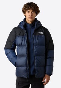 Marinblå pufferjacka med svarta axlar, dragkedjestängning och huva. Tyget har en glänsande yta och har en logotyp på vänster bröst.