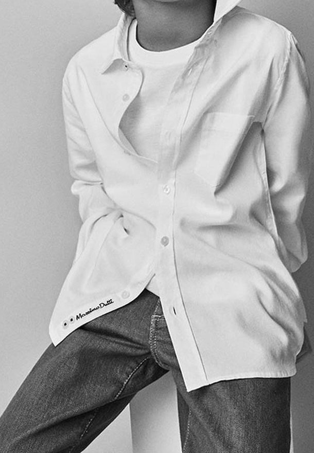 Personne portant une chemise blanche déboutonnée par-dessus un t-shirt blanc et un jean foncé, assise avec les mains dans le dos.