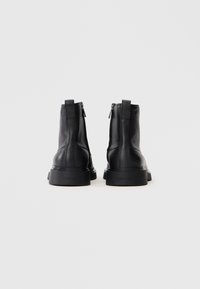 Par de botas de cuero negro hasta el tobillo con cremalleras laterales y tiradores, mostradas desde atrás sobre un fondo blanco liso.
