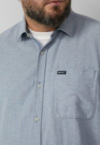 Jasnoniebieska koszula zapinana na guziki, z kieszenią, fakturą tkanej tkaniny oraz małą naszywką z logo na piersi. Guziki mają jasny kolor.