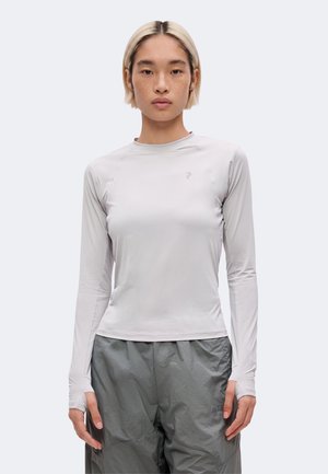Personne aux cheveux blonds courts portant un haut ajusté gris clair à manches longues et un pantalon ample gris foncé, debout devant un fond uni.
